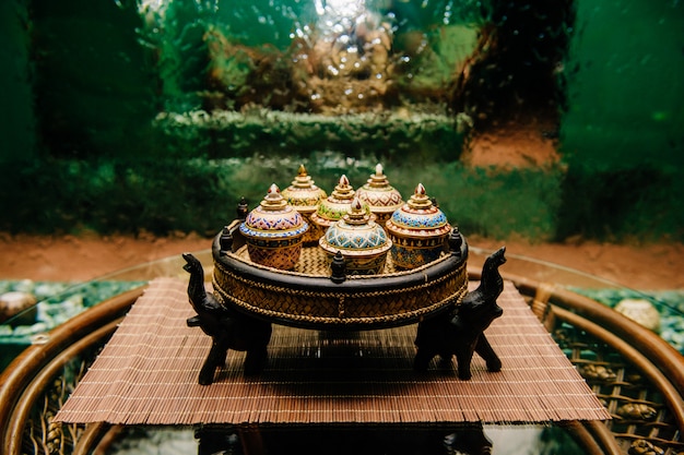 Bouilloire en bronze traditionnelle thaïlandaise célèbre cérémonie sur plateau en osier avec des fleurs de lotus, tasse, sucre et biscuits sur table en rotin avec surface vitreuse avec mur de cascade abstrait sur fond.