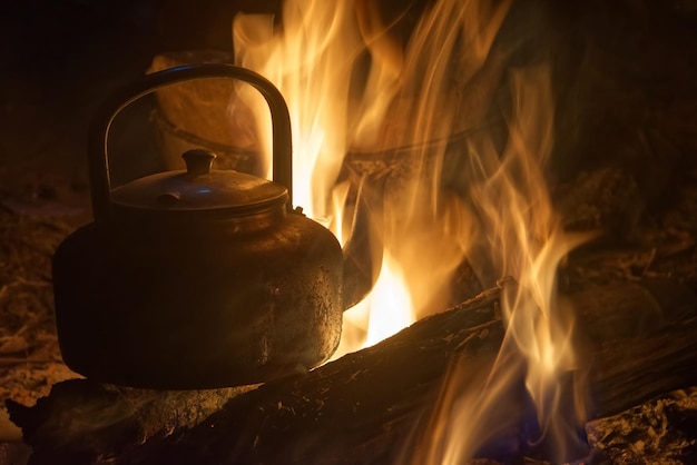 Une bouilloire en métal rouillé sur un tas de bois de chauffage brûlant avec un arrière-plan flou dans un camp