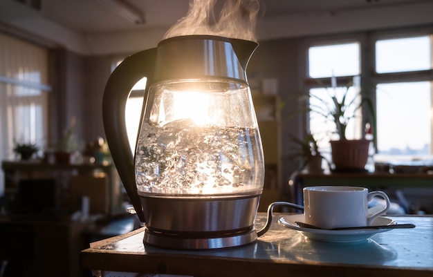 Une bouilloire transparente d'eau bout sur fond de coucher de soleil brillant à travers la fenêtre