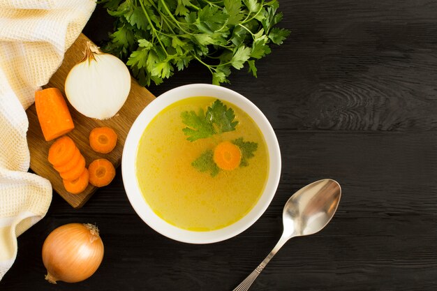 Bouillon de poulet dans le bol blanc et légumes sur la surface en bois noire. Vue de dessus Copiez l'espace.