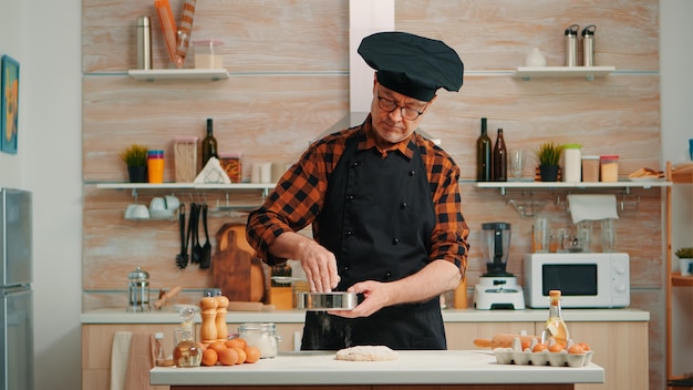 Boulanger avec bonete et tablier utilisant un tamis métallique préparant des produits de boulangerie à la maison. Heureux chef âgé avec uniforme de cuisine mélangeant, saupoudrant, tamisant les ingrédients crus pour faire du pain traditionnel