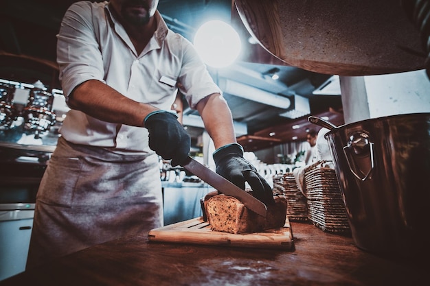 Un boulanger expérimenté dans des gants de protection tranche du pain pour le petit-déjeuner quotidien au restaurant.