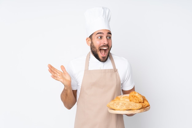 Boulanger mâle tenant une table avec plusieurs pains isolé sur mur blanc avec expression faciale surprise