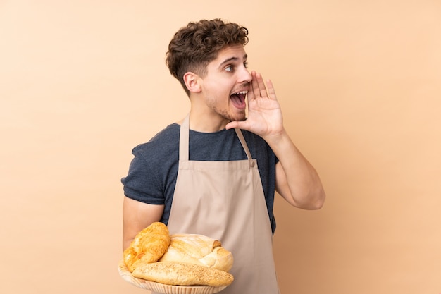 Boulanger mâle tenant une table avec plusieurs pains sur mur beige criant avec la bouche grande ouverte