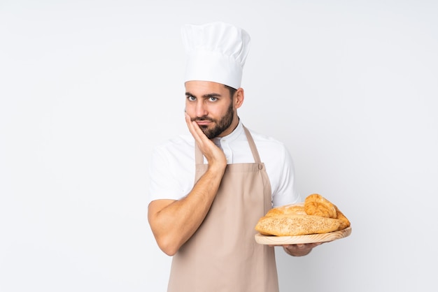 Boulanger mâle tenant une table avec plusieurs pains sur mur blanc malheureux et frustré