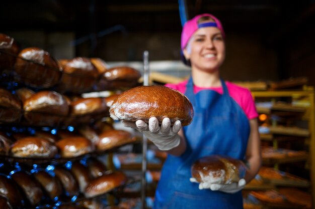 Boulanger Professionnel - Une Jeune Et Jolie Femme Dans Un Tablier En Jean Tient Du Pain Frais Dans Le Contexte D'une Boulangerie Ou D'une Boulangerie. Produits De Boulangerie. Fabrication De Pain