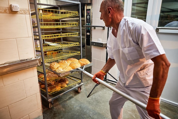 Un boulanger professionnel en uniforme sort un chariot avec du pain fraîchement cuit d'un four industriel dans une boulangerie