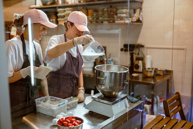 Un boulanger professionnel verse du lait dans un bol en métal sur des balances selon la recette pour faire de la pâte avec un collègue dans une boulangerie artisanale