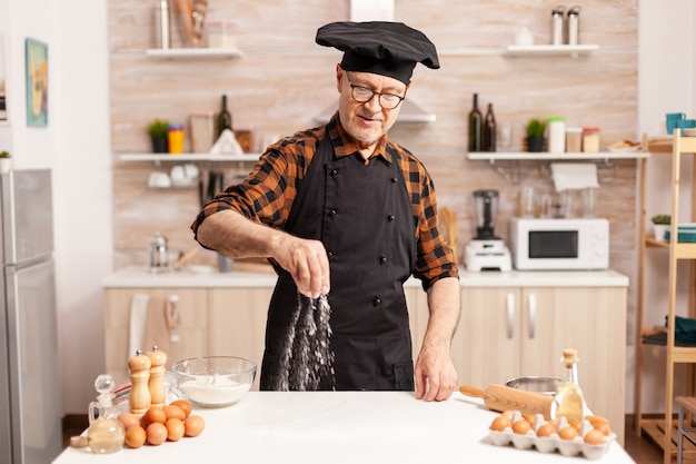 Boulanger retraité portant un tablier et préparant une pizza maison sur la table de la cuisine. Chef senior à la retraite avec bonete et tablier, en uniforme de cuisine saupoudrant tamisage tamisant les ingrédients à la main.