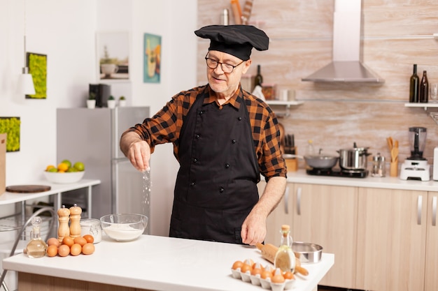 Boulanger senior dans la cuisine à domicile mettant de la farine de blé sur une table en bois. Chef senior à la retraite avec bonete et tablier, en uniforme de cuisine saupoudrant tamisage tamisant les ingrédients à la main.