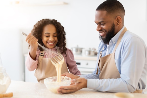 Boulangerie à domicile. Portrait d'une fille afro-américaine souriante mélangeant de la pâte liquide et de la farine dans un bol en verre à l'aide d'un batteur à oeufs, papa assis à table et aidant sa fille à préparer des crêpes dans la cuisine