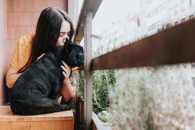 Bouledogue français noir embrassé et câliné par le propriétaire sur la terrasse assis sur un banc en boisChien penché sur un bar regardant la ville à travers des plantesJeune femme heureuse et joyeuse prenant en charge le chiot