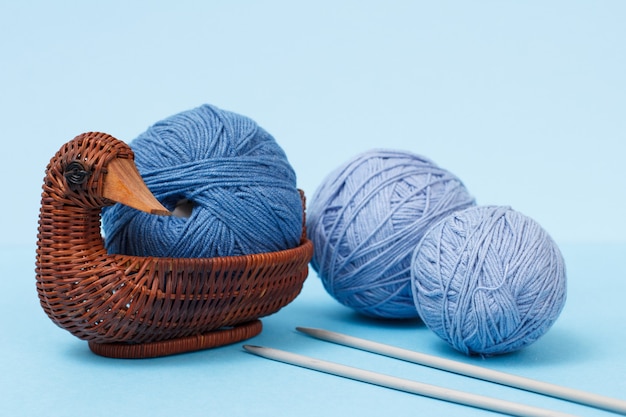 Boules de fil à tricoter, aiguilles à tricoter en métal et panier en osier sur fond bleu. Notion de tricot.