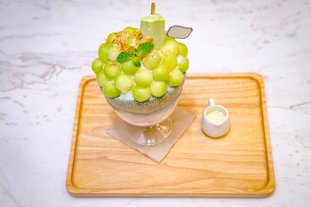 Des boules de melon vert sont disposées sur le dessus du bingsu (style glace coréenne) et décorées avec de la glace au thé vert et une feuille de menthe sur la plaque de bois et du lait concentré sucré à côté.