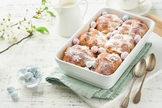 Photo boulets de croix chauds de pâques traits traditionnels de pâque boulettes de croix avec des raisins secs, du beurre, du chocolat, des œufs de bonbons avec des fleurs de cerises ou de pommes en fleurs sur des arrière-plans rustiques en bois clair.