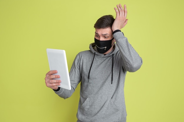 Bouleversé à l'aide de la tablette. Portrait d'homme caucasien isolé sur mur jaune. Modèle masculin bizarre en masque facial noir.