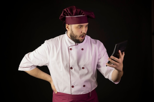 Bouleversé étonné jeune chef barbu en uniforme regardant la calculatrice