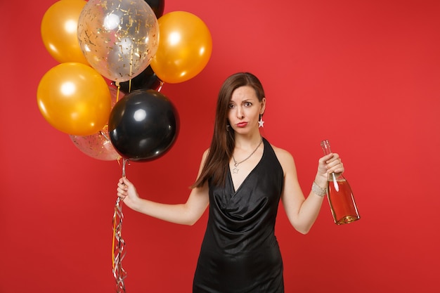 Bouleversé la jeune fille perplexe en petite robe noire célébrant, tenant une bouteille de champagne, des ballons à air isolés sur fond rouge. Saint Valentin, concept de fête de vacances de maquette d'anniversaire de bonne année.