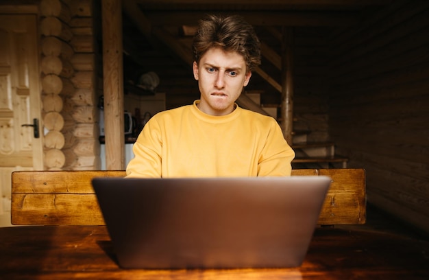 Bouleversé jeune homme assis à une table de maison de campagne avec un intérieur en bois et utilise un ordinateur portable