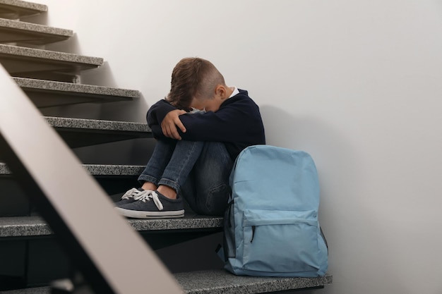 Bouleversé le petit garçon avec le sac à dos se reposant sur des escaliers à l'intérieur