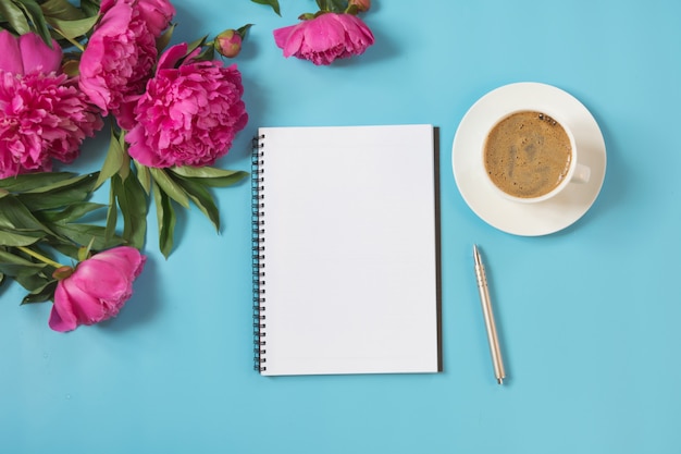 Bouquet de fleurs de pivoine rose, tasse de café pour le petit déjeuner, cahier vide, stylo sur bleu pastel.