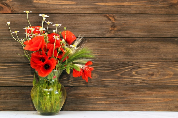 Un bouquet de fleurs sauvages : marguerites et coquelicots dans un vase en verre transparent sur fond de bois.