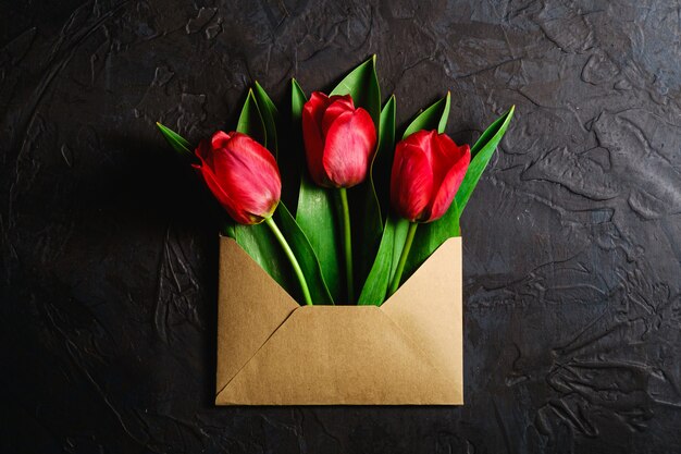 Bouquet de fleurs de tulipe rouge dans une enveloppe en papier sur fond noir foncé texturé, vue de dessus copie espace