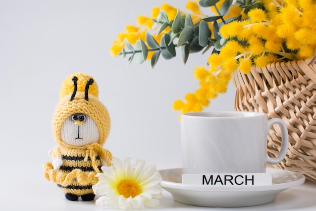 Un bouquet jaune de mimosa et une tasse avec une boisson l'inscription mars et une abeille tricotée jaune sur fond blanc Le concept des vacances de printemps le 8 mars carte de voeux
