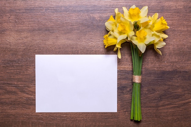 Un bouquet de jonquilles et une feuille de papier blanc sur un fond en bois. Fleurs jaunes de printemps. Papier avec espace pour le texte. Conception à plat, vue de dessus.