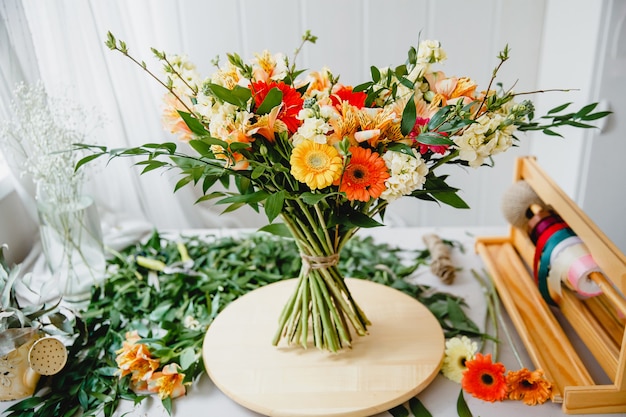 Un bouquet luxuriant dans le style bohème sur un support en bois sur une table jonchée de feuilles et d'outils dans l'atelier du fleuriste