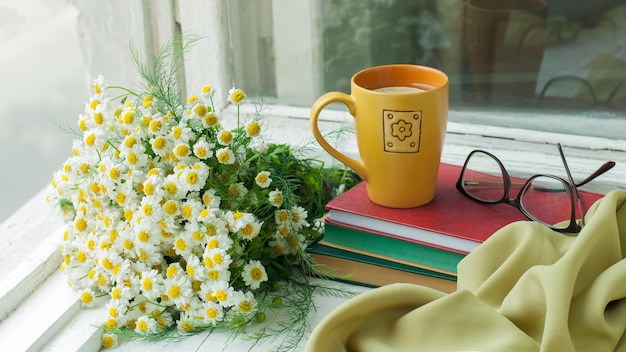 Un bouquet de marguerites une tasse de thé et une pile de livres sur un rebord de fenêtre en bois un jour d'été