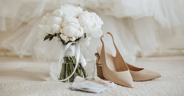 Bouquet de mariage de fleurs de pivoines dans un vase se dresse sur le lit des jeunes mariés avec des invitations et des chaussures sur l'arrière-plan