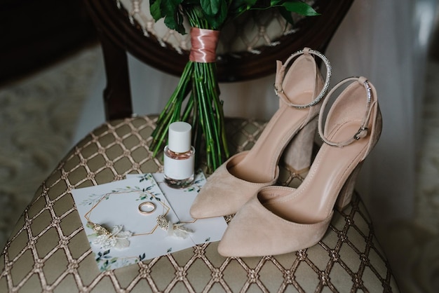 Bouquet de mariage de la mariée chaussures beiges élégantes parfum boucles d'oreilles et bague sur fond de chaise Accessoires de mariée Lettres de la mariée et du marié Vue latérale