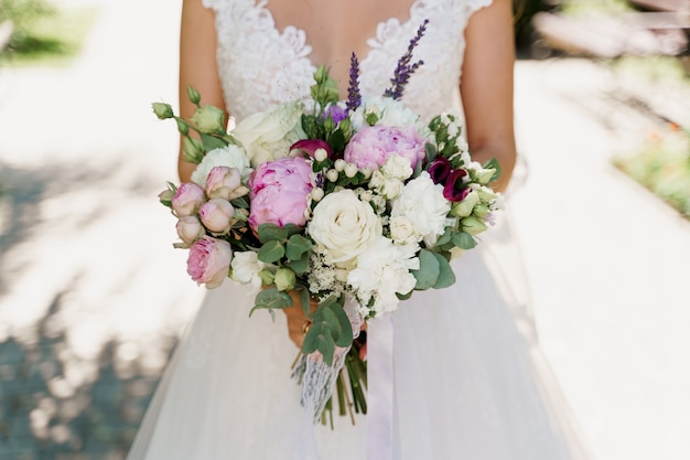 Bouquet de mariage avec des roses blanches, des pivoines et des feuilles vertes. La mariée en robe tient le bouquet.