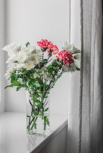 Bouquet d'oeillets de chrysanthèmes dans un vase en verre sur le rebord de la fenêtre dans une pièce confortable