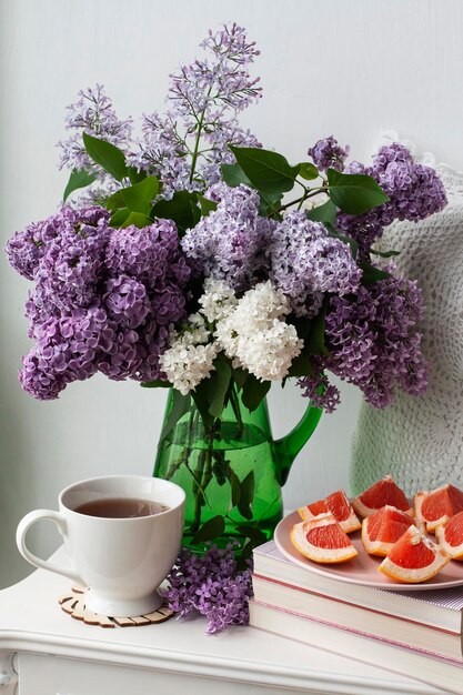 Un bouquet parfumé de lilas colorés se dresse dans un vase en verre vert sur fond blanc à côté d'une tasse de thé chaud et de tranches de pamplemousse