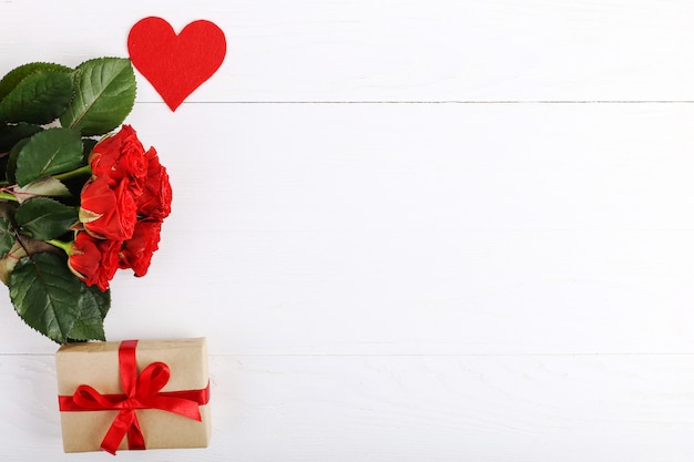 Un bouquet de roses rouges, un cadeau et des coeurs sur une table en bois blanche