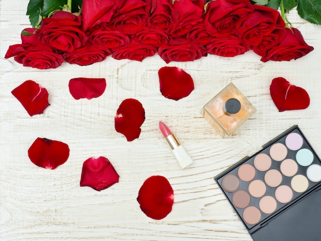 Bouquet de roses rouges, palette de parfum, rouge à lèvres et fard à paupières sur une table en bois blanche.