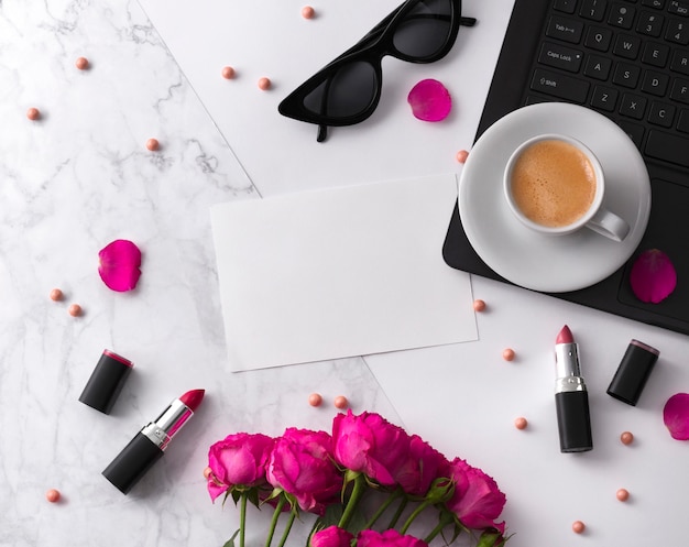 Bouquet de roses, tasse de café, ordinateur portable, lunettes de soleil et rouge à lèvres sur un tableau blanc.