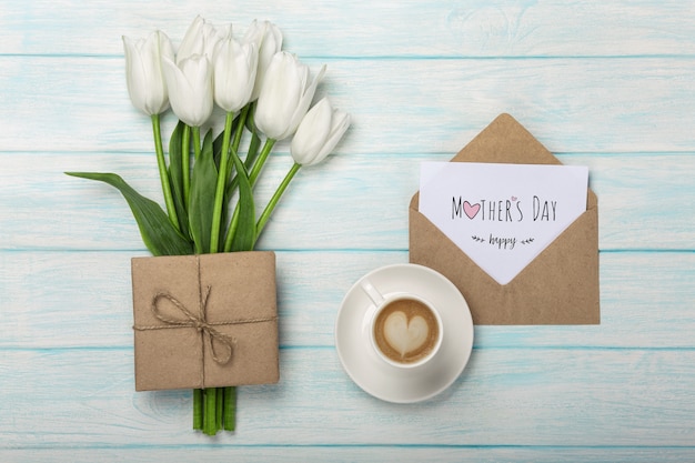 Un bouquet de tulipes blanches, une tasse de café avec une note d&#39;amour et une enveloppe sur des planches en bois bleues. Fête des mères