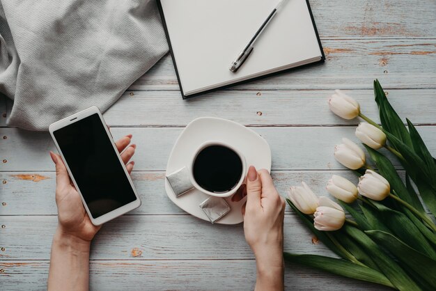 Bouquet de tulipes blanches avec une tasse de café et un smartphone dans la main de la femme et un cahier vide sur une table en bois