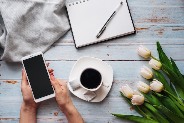 Bouquet de tulipes blanches avec une tasse de café et un smartphone à la main des femmes et un cahier vide sur une table en bois
