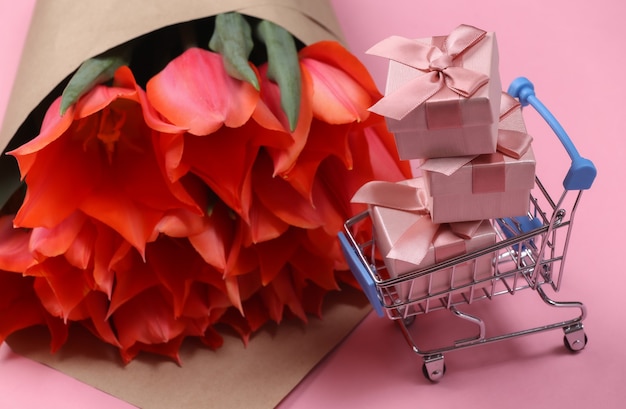 Bouquet de tulipes et caddie avec coffrets cadeaux sur fond rose. Achats de vacances. Fête des mères ou 8 mars, anniversaire.
