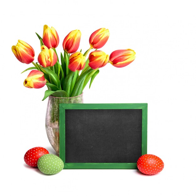 Bouquet de tulipes rouges avec des sommets jaunes, un tableau noir et des oeufs de Pâques colorés sur blanc, espace de texte