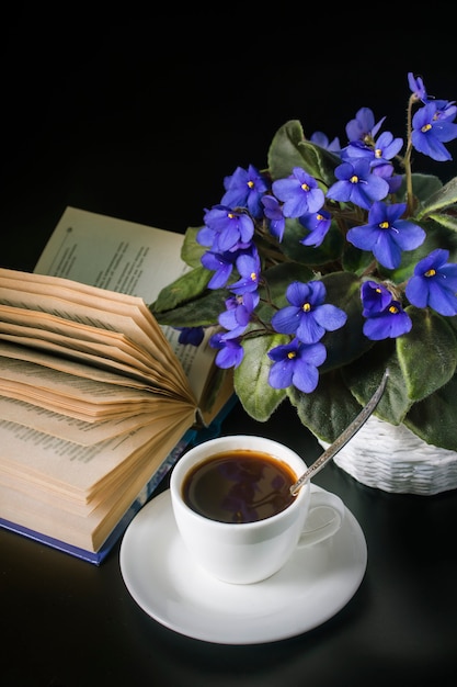 Bouquet De Violettes Africaines Avec Une Tasse De Café Et Un Livre Ouvert Sur Une Surface Noire.