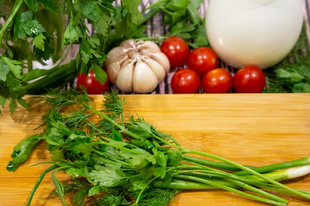 Bouquets d'oignons verts, de persil et d'aneth sur une planche à découper en bois à côté de tomates, pommes de terre, ail et crème sure. Ingrédients pour la salade