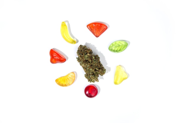 Un bourgeon sec de marijuana se trouve sur un fond blanc entouré de bonbons gommeux à mâcher de différentes formes et couleurs sur fond blanc Vue de dessus beaucoup d'espace vide