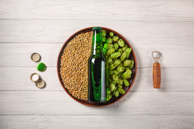 Une bouteille de bière sur une assiette avec houblon vert et grain d&#39;avoine avec ouvre-porte et corcks sur une planche de bois blanche