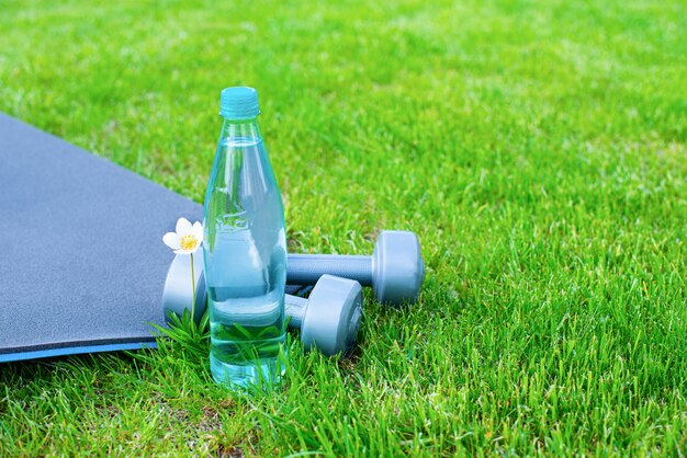 Bouteille d'eau, haltères et un tapis pour le sport et le yoga sur l'herbe verte. Espace copie