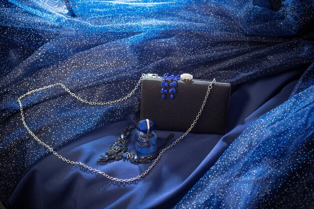 Bouteille de parfum pochette noire et bijoux sur fond de robe de soirée bleue